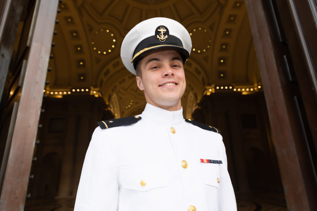 Naval Academy graduate in white uniform smiles at Bancroft Hall.