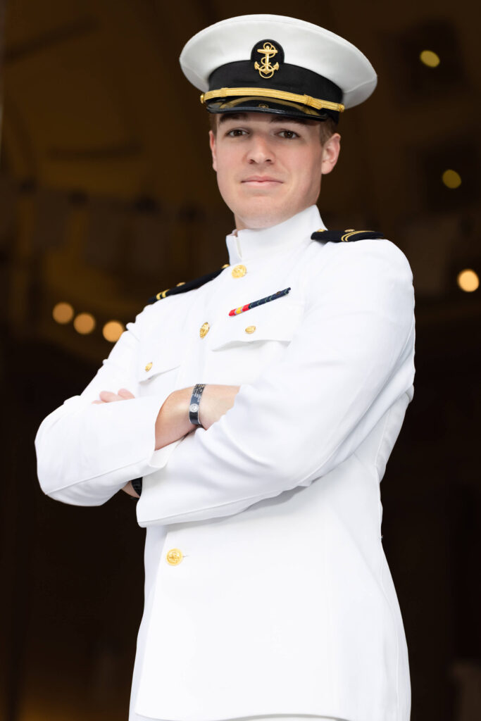 Naval Academy graduate in white uniform smiles at Bancroft Hall.