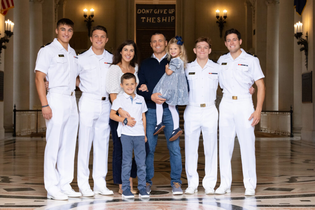 Sponsor Family of 4 and Four USNA Midshipmen in Summer White Uniform stand in the Rotunda of Bancroft hall and smile at camera during a photo shoot with Kelly Eskelsen Photography in Annapolis Maryland.